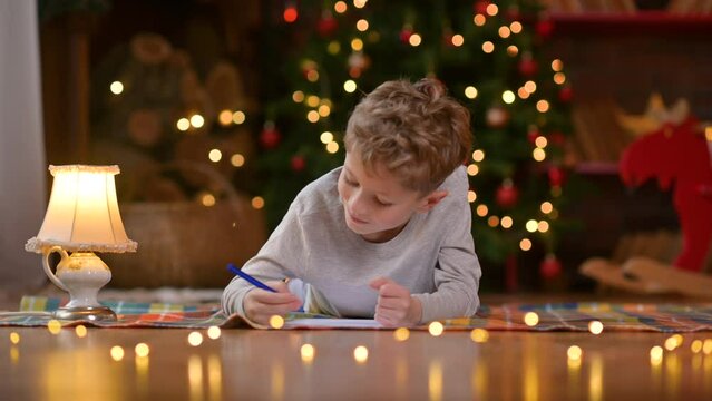 Portrait of a little boy lies on the floor and writes a letter to Santa Claus against the background of a festive Christmas tree. holidays and celebrations concept