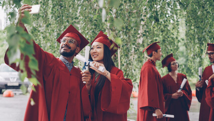 Obraz premium Two students are taking selfie with graduation diplomas wearing mortarboards and gowns, guy is holding smartphone and taking picture, girl is posing.