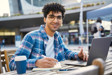 Smiling handsome middle eastern student studying using laptop computer, online education concept. Portrait of happy successful Egyptian programmer working freelance project online at workplace