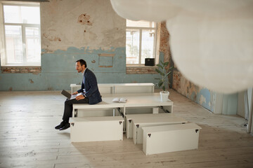 Portrait of young businessman sitting in empty room and working on laptop on new business project....