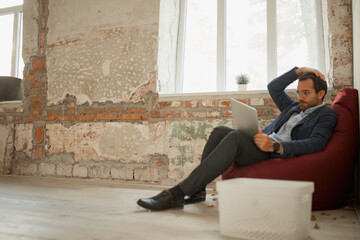 Portrait of young businessman with thoughtful expression sitting on floor pillow with laptop and working. Business closure. In search of vacancies