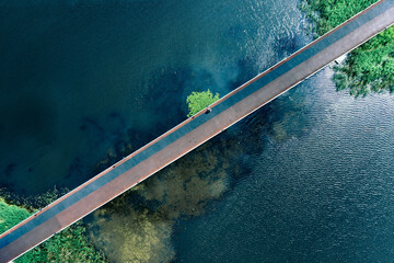 Top view of footbridge across amazing blue ripples water texture pond. Drone view of city bridge over lake, summer cityscape background. Diagonal bridge with sports running rubber coating.