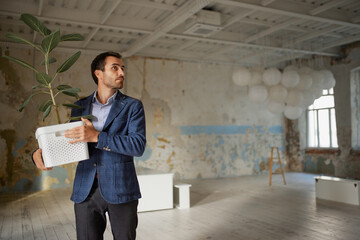 Portrait of young businessman walking out empty room with flower. Business closure. Depression and crisis