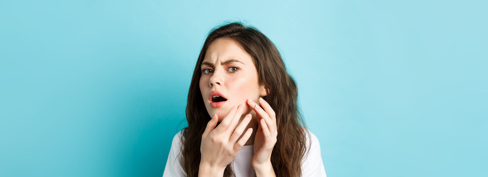 Skin Care. Image Of Worried Teenage Girl Looking In Mirror And Spot A Pimple On Face, Trying Squeeze Acne On Cheek, Frowning Upset, Standing Over Blue Background