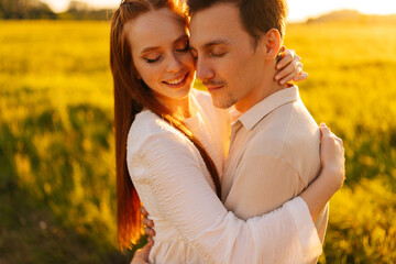 Close-up faces of young couple in love with closed eyes standing hugging together on beautiful green meadow in summer evening, during sunset with soft sunlight. Concept of romantic date love outdoors.