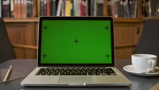 Close up footage of silver macBook pro laptop computer screen display with green screen which is good for mock ups. Laptop is placed on the table with a library or book store background.