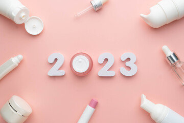 Best skincare products and cosmetic trends of 2023 concept. 2023 white number with lip balm, cream bottle, serum and lotion on pink background. - obrazy, fototapety, plakaty
