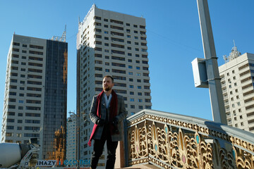 Stylish young man with a beard in a gray coat, red boots and gloves stands on the stairs in the city. In the background, the city, buildings, blue sky. Style, fashion, gentleman.