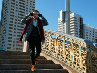 Stylish young man with a beard in a gray coat, red boots and gloves stands on the stairs in the city. Headphones. In the background, the city, buildings, blue sky. Style, fashion, gentleman.