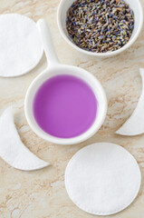 Small white bowl with purple lavender water (toner, micellar water) and cotton pads. Ingredients for preparing homemade cosmetics. Natural beauty treatment recipe.