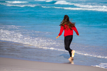 Back view of a long-haired girl running along the ocean shore on a paradise beach at sunset;...