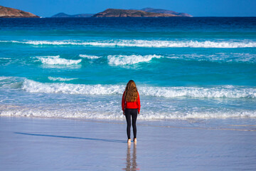 A back view of a long-haired girl standing by the ocean shore on a paradise beach at sunset;...