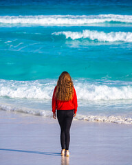 Fototapeta na wymiar A back view of a long-haired girl standing by the ocean shore on a paradise beach at sunset; turquoise water on a beach in lucky bay, western australia