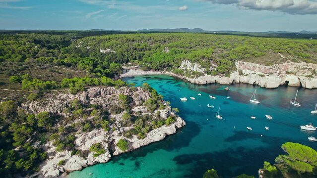 Aerial drone view of people having a good time by the beach with turquoise water. Tourists enjoy Cala Macarelleta and Macarella on the Balearic Islands, Spain, swimming and sailing yachts and boats.
