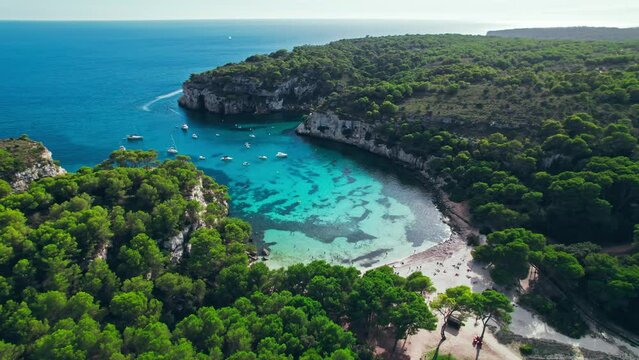 Aerial drone view of people having a good time by the beach with turquoise water. Tourists enjoy Cala Macarelleta and Macarella on the Balearic Islands, Spain, swimming and sailing yachts and boats.