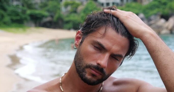 Attractive young dark-haired man, bearded macho staring to the camera. Confident middle-aged Caucasian guy with naked torso, gazing with an insightful look, enjoying sunbathing on the tropical beach