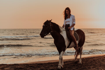 Woman in summer clothes enjoys riding a horse on a beautiful sandy beach at sunset. Selective focus 