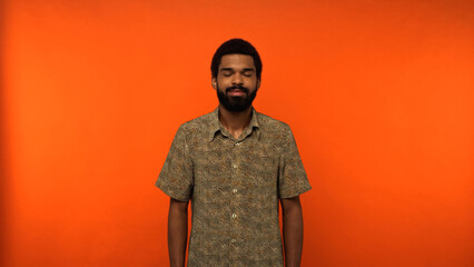bearded and pleased african american man with closed eyes standing in shirt on orange background.