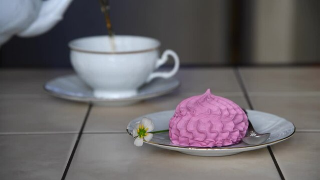 Pink meringue in white saucer lies on table with spoon and one flower. Blurred person pouring tea from teapot in white cup in the background. Selective focus. Real time video. Tea time theme.