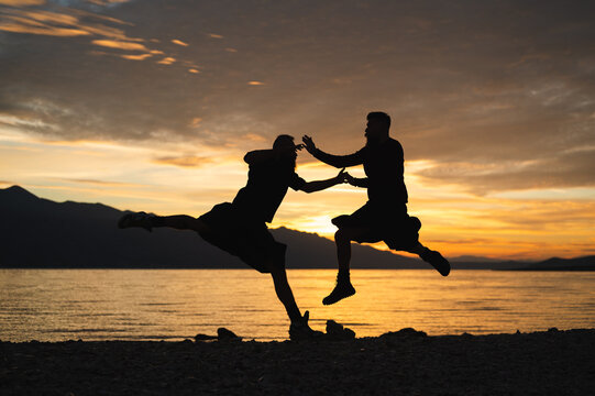 Gay couple in kilts jumping on the beach in sunrise. Silhouette of homosexual partners having early morning fun by seaside.