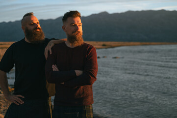 Attractive hipster long beard gay couple enjoying sunrise on beach, sunbathing on morning sun. Romantic male homosexual partners outdoors during golden hour by seaside.