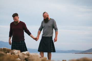 Fashionable gay couple in kilts holding hands while walking by sea side. Long beard male homosexuals enjoying on the beach, overcast day.