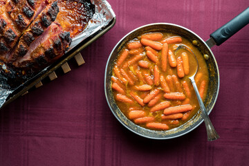 Sauteed carrots in a fry pan with a glazed smoked picnic ham in a pan beside.