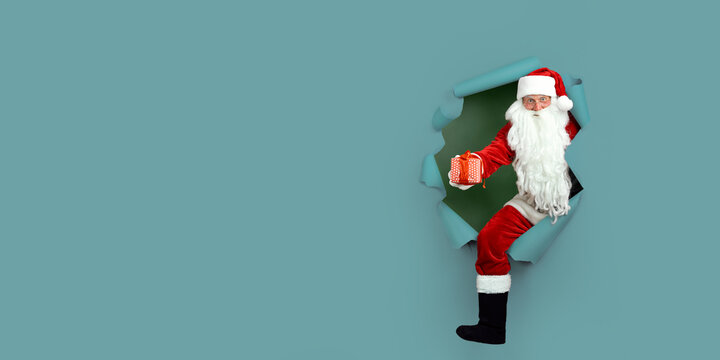 Full length of Santa Claus breaks through hole in paper with giftbox. Holiday poster design, banner. - Powered by Adobe