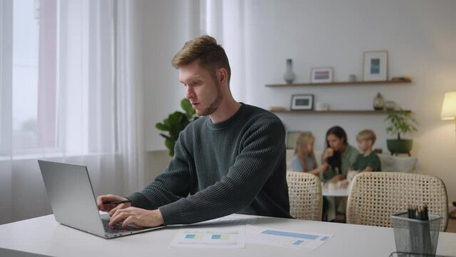 Mid 30s Caucasian male working from home, having a video call with colleagues. Family playing in the background.