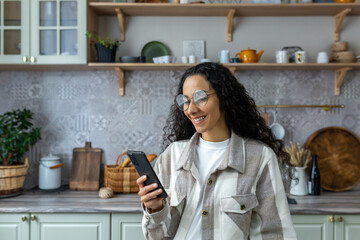 Hispanic woman uses mobile phone at home in the kitchen, woman smiles and rejoices, reads messages, browses web pages, online communication with friends and dating.