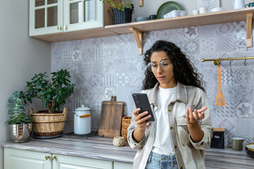 Sad hispanic woman in depression reading bad news on smartphone, woman in glasses and curly hair using smartphone at home in kitchen.