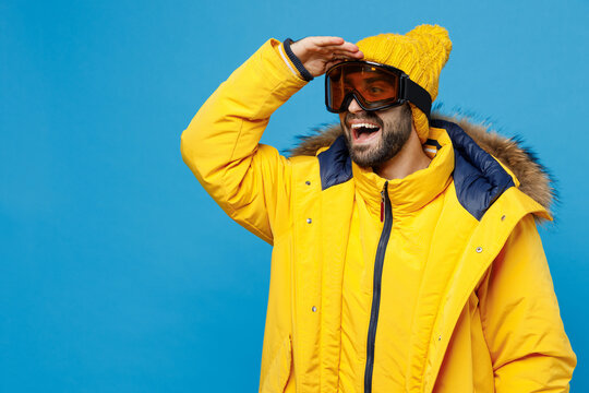 Young man 20s wears ski goggles yellow puffer down jacket with fur hood hold hand at forehead look far away distance isolated on plain blue background studio portrait. People winter lifestyle concept.