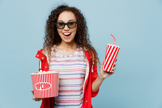 Young excited woman of African American ethnicity in 3d glasses watch movie film hold bucket of popcorn cup of soda pop in cinema isolated on plain pastel light blue cyan background studio portrait.