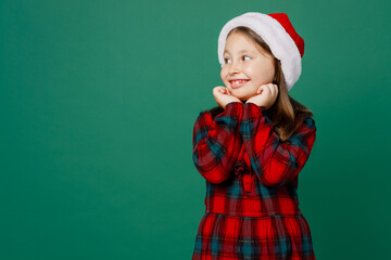 Merry smiling happy little child kid girl 6-7 years old wear red dress Christmas hat posing hold face look aside on workspace area isolated on plain dark green background. Happy New Year 2023 concept.