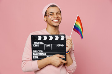 Young gay man wear sweatshirt hat hold striped rainbow flag classic black film making clapperboard isolated on plain pastel light pink color background studio portrait. Lifestyle lgbtq pride concept.