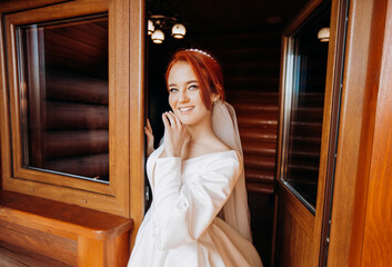 Amazing red-haired bride in a satin white dress is posing on the balcony on the background of a room in a wooden style.