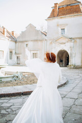 Amazing red-haired bride in a satin white dress with a veil poses in the courtyard of an ancient castle.