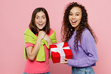 Young two friends shocked excited surprised women 20s wears green purple shirts together hold red present box with gift ribbon bow isolated on pastel plain light pink color background studio portrait.