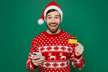 Merry young man wear red warm sweater Santa hat posing use mobile cell phone credit bank card...
