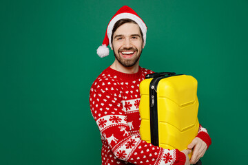 Traveler man wear knitted sweater Santa hat hold valise isolated on plain dark green background. Tourist travel abroad in free spare time rest getaway. Air flight trip journey concept New Year 2023.
