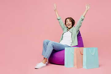 Full body young woman wears green shirt sit in bag chair near paper package bags after shopping raise up hands isolated on plain pastel light pink background studio. Black Friday sale buy day concept.