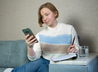 girl sits on sofa with phone and notebook and studies online