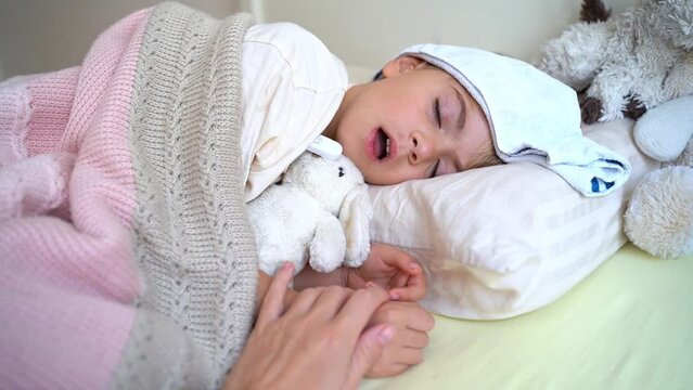 Mom hand holds hand of sick fever cough child lying in bed with cold compress on his forehead, measure body temperature with electronic thermometer. Children cold and flu, coronavirus illness concept.