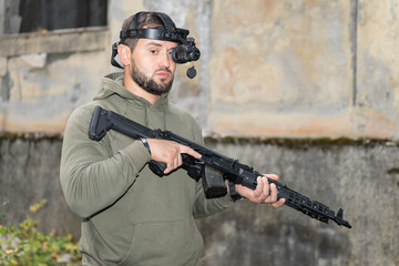 Fototapeta premium A man with a night vision device and a rifle in his hands near an abandoned building.