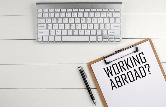 Home office workspace with keyboard, clipboard and pen with text WORKING ABROAD on white wooden background , business concept