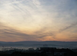 Sonnenaufgang über Windmühlen, Wald und Häuserdächern in Stadt am frühen Morgen bei Nebel im Herbst