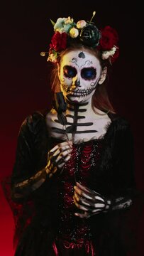 Vertical video: Flirty woman wearing santa muerte body art in studio, acting glamorous on day of the dead mexican halloween celebration. Ritual skull body art, posing with black roses and flowers