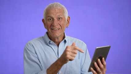 Animated elderly pointing at digital tablet computer screen with finger. Retired old man spend time talking to friends and family isolated on solid purple background