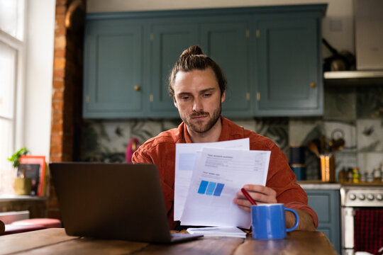 Young adult male at home looking at bills and personal finances using a laptop