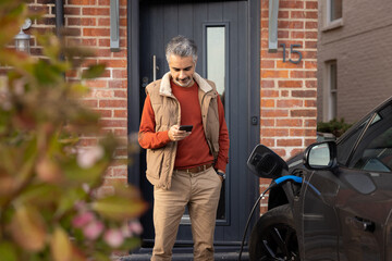 Mid adult male charging electric car and using smartphone app at home